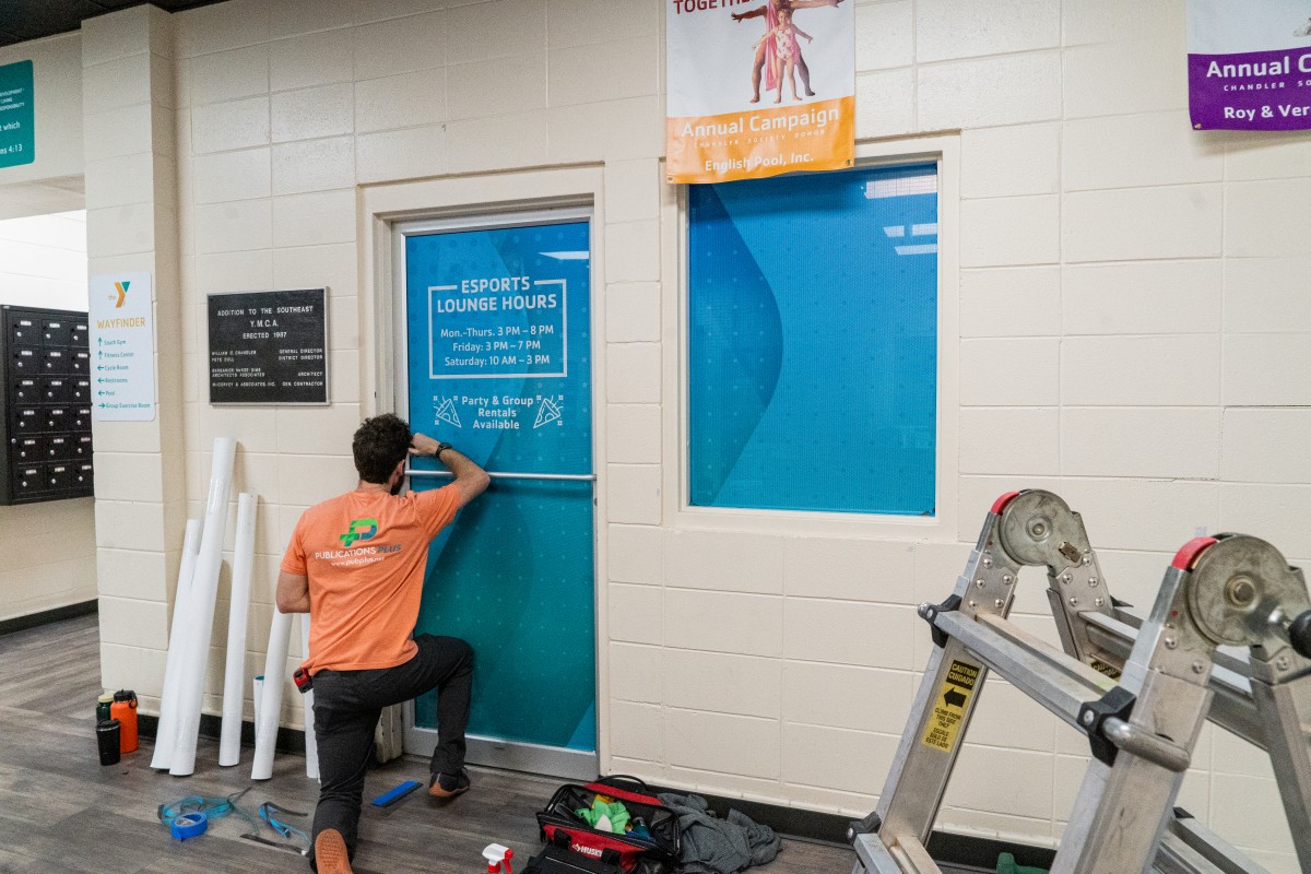 Two men working on installing a wall wrap decal, one on a ladder, the other on foot.